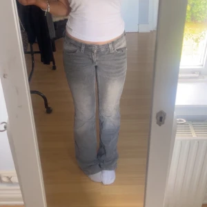 Grå bootcut jeans med broderade fickor - Säljer ett par gråa bootcut jeans och snygga broderade detaljer på bakfickorna. Jeansen har låg midja och är i klassisk denim med slitningar nertill. Den ena knappen på baksidan är borta tyvärr men går att fixa lätt om man har knapp, det är också ett hål längst nere på vänstra benet men man ser inte det när man går/står