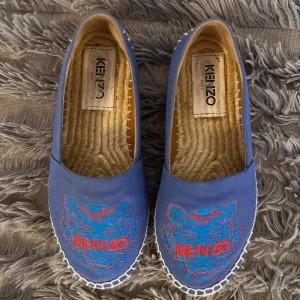 Blå espadrillos från Kenzo - Snygga blå espadrillos från Kenzo med ikoniskt broderat tigerhuvud i blått och rött framtill. Skorna har en klassisk rund tå, platt sula i rep och ovandel i bomull. Perfekta för sommaren och knappt använda. De är i gott skick! 