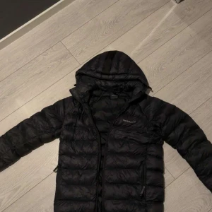 Svart Peak Performance pufferjacka - Svart pufferjacka från Peak Performance med huva och dragkedja framtill. Jackan har quiltad design, två sidofickor med dragkedja och ribbade muddar vid ärmsluten. Perfekt för kalla dagar och riktigt snygg streetstil.🥰 jätte fint skick! kan gå med på att sänka lite.