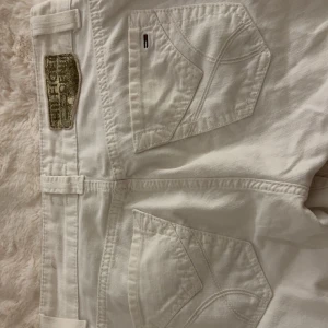 Vita jeans från Tommy Hilfiger - Säljer ett par vita jeans från Tommy Hilfiger med klassiska bakfickor och snygga detaljer. Byxorna har dragkedja, knapp med logga och breda hällor. Materialet är bomull och de har en rak passform. Perfekta för dig som gillar stilrena och fräscha jeans.