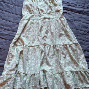 Grön blommig klänning med smock, M - Supersöt klänning i ljusgrönt med vita blommor, smockad upptill och tunna axelband. Klänningen har volangdetaljer och är perfekt för varma dagar. Materialet känns lätt och luftigt, troligtvis bomull eller polyester. Justerbara axelband för bästa passform.