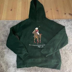 Grön hoodie från Ralph Laurent  - Säljer en snygg grön hoodie från Ralph Lauren med en cool Polo Bear-design på framsidan. Perfekt för höst och vinter med sin mysiga känsla. Den har en klassisk passform och är i mycket bra skick. Perfekt för att hålla stilen både i skolan och på fritiden! 🍂🐻