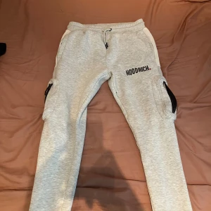 Grå mjukisbyxor från Hoodrich M - Snygga ljusgrå mjukisbyxor från Hoodrich med svarta och röda tryckdetaljer. Byxorna har snörning i midjan, cargofickor med dragkedja på sidorna och en relaxed passform. Perfekta för chill dagar eller streetwear-stil.