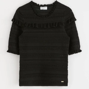 Svart ribbad tröja med volang Holly & Whyte - Supersnygg svart ribbad tröja från Holly & Whyte med volangdetaljer vid halsringning och axlar. Tröjan har lång ärm och är stickad i ett mjukt material. Använd en gång!!