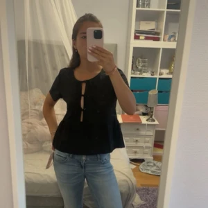 Svart blus med knyt framtill - Säljer en svart blus med kort ärm och tre dekorativa knyt framtill. Blusen har en lätt utsvängd passform och rundad halsringning. Perfekt för dig som gillar trendiga detaljer och enkel stil.