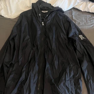 Svart vindjacka från Risky med huva - Svart vindjacka från Risky med huva och coolt tryck med droppande R på ärmen och huvan. Jackan har dragkedja framtill och är tillverkad i lätt syntetmaterial, perfekt för blåsiga dagar. Passformen är loose och jackan har långa ärmar.