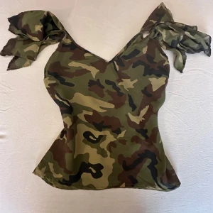 Camouflage topp med knytband - Säljer en snygg camouflagefärgad topp med knytband på axlarna. Toppen har v-ringad hals och är ärmlös, perfekt för dig som gillar statement-plagg. Materialet känns lätt och smidigt, och mönstret går i grönt, brunt och svart.