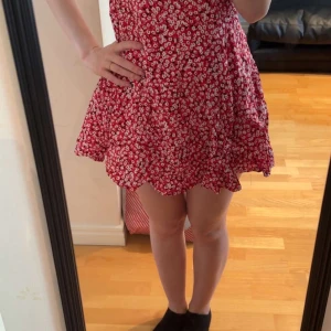 Röd blommig kortklänning  - Supersöt röd kortklänning från Ambar med vita blommor och knytband i ryggen. Klänningen har en flowy passform och är perfekt för varma dagar. Tillverkad i viskos som känns lätt och skön mot huden. Perfekt för dig som gillar färg och mönster!