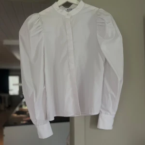 Vit skjorta med puffärm från NA-KD - Säljer en stilren vit skjorta från NA-KD med puffiga axlar och lång ärm. Skjortan har dold knäppning framtill och klassisk krage. Perfekt för dig som vill ha en clean look med lite extra volym på ärmarna.