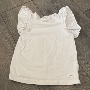 Vit blus med volangärm från Newbie - Supersöt vit blus från Newbie i mjuk bomull. Toppen har korta volangärmar med spetsdetaljer och rund halsringning med söt kant. Perfekt för en gullig och stilren look till de minsta.
