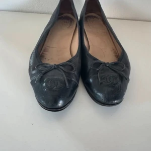 Chanel ballerina - Klassiska svarta ballerinas från Chanel i mjukt skinn med rund tå, dekorativ rosett framtill och ikoniska CC-loggan broderad på tån. Snygg och tidlös modell som passar till allt. Sula i läder och diskret Chanel-stämpel undertill.