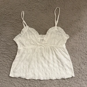Vit topp - Säljer denna supersöta linnetopp med tunna axelband och broderade blommor. Toppen har en liten rosett framtill som man kan justera och vågig kant vid bysten. Perfekt för varma dagar och ger en romantisk vibe. Lätt och luftig känsla med fina detaljer.