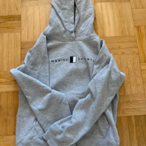 Grå hoodie från Marine Sports L - Grå hoodie från Marine Sports med tryck på bröstet. Klassisk modell med huva och dragsko, samt känguruficka framtill. Mjuk insida och normal passform, perfekt för chill dagar. Skandinavisk sportswear-vibe💗