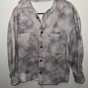 Grå overshirt med batikmönster - Säljer en oversized grå overshirt med batikmönster. Skjortan har två stora bröstfickor, knappar framtill och klassisk krage. Perfekt för lager-på-lager och chill stil. Materialet känns som bomull och ger en avslappnad vibe.
