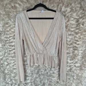 Guldbeige omlottopp Gina Tricot - Säljer en glittrig guldbeige omlottopp från Gina Tricot. Toppen har lång ärm, v-ringning, volangkant nedtill och är sydd i ett plisserat, skimrande material som ger en lyxig känsla. 