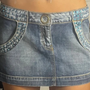 Jeanskjol med blå nitar och detaljer - Snygg kort jeanskjol med coola blå nitar längs fickorna och midjan. Kjolen har en markerad guldfärgad knapp och dekorativa sömmar. Klassisk femficksmodell med slitningar och en unik look som sticker ut.