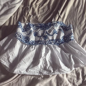 Vit och blå broderad linnetopp H&M - Supersöt vit linnetopp från H&M med tunna axelband och blått broderat blommönster över bysten. Toppen har en volangig, luftig passform och smock i ryggen för extra komfort. Perfekt för varma dagar och ger en fräsch sommarkänsla.