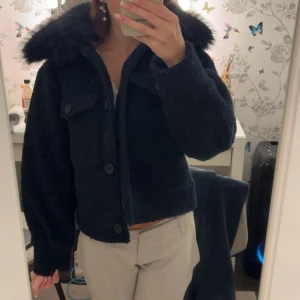 Mörkblå teddyjacka med fuskpäls - Mysig mörkblå teddyjacka med stor krage i fuskpäls och stora knappar framtill. Jackan har en croppad passform och en ficka med lock på bröstet. Perfekt för kyliga dagar när du vill vara både varm och snygg. Storlek 158/164, jätte fint skick (aldrig använd)