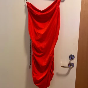 Röd kort klänning med kedjedetaljer - Säljer en riktigt snygg röd kortklänning med draperad passform och kedjor som axelband och detalj bak. Klänningen har en cool rynkad effekt och är ärmlös med en trendig look. Materialet känns som polyester. Pris kan diskuteras vid snabb köp 