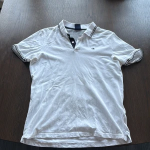 Vit pikétröja från Champion M - Vit pikétröja från Champion med svart broderad logga på bröstet. Klassisk krage med svarta ränder och knäppning framtill. Ärmarna har matchande svarta ränder och tröjan är gjord i bomull för en skön känsla. Perfekt för en clean och sportig stil.