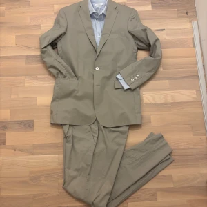 J.Lindeberg kostym - Snygg beige kostym från J.Lindeberg i klassisk modell med en modern twist. Kavajen har två knappar, fickor med lock och lätt vadderade axlar. Tillverkad i 90 % bomull och 10 % polyester för en lätt och bekväm känsla. Designad med en något sportigare känsla än traditionella kostymer, gjord för golfbanan. Kavaj i storlek 50, byxa i storlek 48. Hör av dig vid frågor 😊