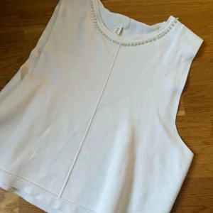 Vit ärmlös topp med pärldetaljer H&M - Säljer en vit ärmlös topp från H&M med rund halsringning dekorerad med pärlor. Toppen har en rak passform och är tillverkad i ett mjukt, stickat material. Perfekt för att lyfta en enkel outfit med lite extra detalj.