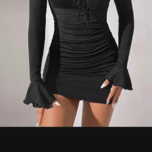 Svart kort klänning med volangärm - Supersnygg svart kort klänning med långa ärmar och volang vid ärmsluten. Klänningen har åtsittande passform, rynkade detaljer och knytning vid bysten. Perfekt för dig som vill sticka ut med en trendig look.