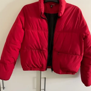 Röd pufferjacka från Divided, stl S - Riktigt snygg röd pufferjacka från Divided med hög krage och dragkedja framtill. Jackan har två fickor med dragkedja och elastisk nederkant för en schysst passform. Perfekt för kalla dagar när du vill sticka ut lite extra.