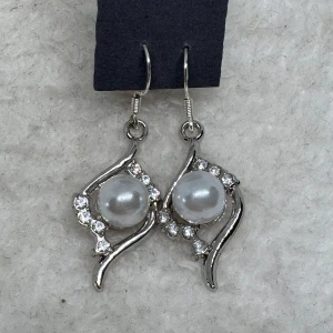 Silvriga örhängen med pärlor - Snygga silvriga örhängen med rund pärla i mitten och gnistrande stenar runt om. Designen är böljande och ger ett elegant intryck, perfekt för dig som gillar att sticka ut med dina accessoarer. Hängande modell som ger extra bling till din look. Krokarna är i sterling silver, nya