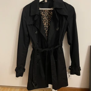 Svart trenchcoat med leopardfoder - Stilren svart trenchcoat med knytskärp i midjan och dubbelknäppning. Jackan har långa ärmar med spänndetaljer vid handlederna och ett snyggt leopardmönstrat innerfoder. Perfekt för dig som vill ha en klassisk look med en twist.