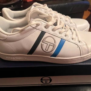 Vita sneakers från Sergio Tacchini - Snygga vita sneakers från Sergio Tacchini med klassisk logga på sidan och svarta samt blå detaljer. Skorna har snörning och är tillverkade i syntetmaterial med en platt sula. Perfekta för dig som gillar stilrena och sportiga skor med en touch av färg.