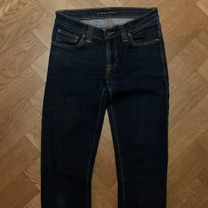 Mörkblå jeans från Nudie Jeans, W27 L34 - Snygga mörkblå jeans från Nudie Jeans med klassisk femficksdesign och kontrastsömmar. Modellen har raka ben och ikonisk broderad detalj på bakfickorna. Tillverkade i slitstarkt bomullsjeansmaterial och passar perfekt till en avslappnad stil.
