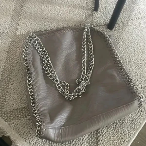 Grå axelväska med kedjedetaljer Tiamo - Snygg grå axelväska från Tiamo i skinnimitation med grova silverfärgade kedjor som axelrem och dekor runt kanterna. Väskan har dragkedja baktill och ett rymligt svart foder med innerfack. Perfekt för dig som gillar en cool och trendig look. Lite slitet men syns typ knappt