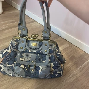 Blommig handväska från River Island - Snygg handväska från River Island med blått och grått blommönster. Väskan har detaljer i grått skinn och mässingsfärgade metalldelar. Praktisk ficka framtill med spänne och dubbla handtag. Perfekt för dig som gillar unik och färgglad stil.