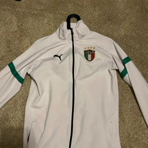 Italien fotbollströja Puma vit S - Snygg vit långärmad fotbollströja från Puma med Italiens landslagsemblem på bröstet och svarta detaljer. Dragkedja framtill och gröna ränder på ärmarna. Perfekt för dig som vill visa ditt stöd för Italien. Materialet är polyester.