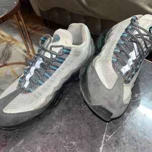 Nike Air Max 95 grå/blå sneakers - Nike Air Max 95 sneakers i grått, vitt och ljusblått med svarta detaljer. Skorna har en ikonisk vågig design, mesh och mocka-paneler, synlig Air-sula och snörning framtill. Perfekta för dig som gillar klassiska och sportiga sneakers med retrovibe.