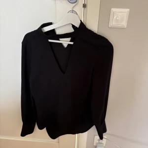 Svart blus med v-ringning från Zara - Svart blus från Zara Woman i storlek XS med lång ärm och snygg v-ringning framtill. Blusen har en unik detalj vid halsen med en öppen del och hög krage. Perfekt för dig som gillar stilrena och trendiga plagg.