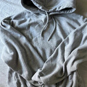 Grå hoodie med huva och snörning - En klassisk grå hoodie med huva och snörning. Tröjan har långa ärmar och ribbade muddar vid ärmslut och nederkant. Perfekt för en avslappnad och chill stil.
