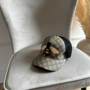 Gucci keps med bi och GG-mönster - Snygg keps från Gucci med klassiskt GG-mönster i beige och brunt, stor bi-tryck framtill och svart mesh baktill. Perfekt statement-piece för dig som vill sticka ut. Justerbar passform och exklusiv känsla med märkets ikoniska detaljer.