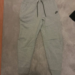 Grå mjukisbyxor från Nike, storlek M - Gråa mjukisbyxor från Nike med svart logga på vänster ben. Byxorna har svarta snören i midjan, ribbade muddar vid bensluten och praktiska fickor med dragkedja. Perfekta för chill eller träning, tillverkade i mjuk bomullsblandning. Inga skador och knappt använda