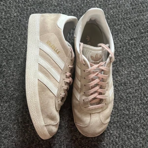 Beige/ljusrosa Adidas Gazelle sneakers strl 36 - Säljer ett par snygga Adidas Gazelle sneakers i beige-rosa mocka med vita detaljer och klassiska tre ränder på sidorna. Skorna har ljusrosa snören och vit sula. Perfekta för dig som gillar stilrena och tidlösa sneakers med en twist. Säljer då de blivit försmå. Använda en säsong, lite grå i kanterna men annars bra skick. Priset går att diskutera. 
