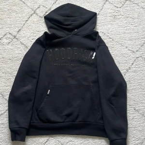 Svart hoodie från Hoodrich - Svart hoodie från Hoodrich med stor logga i relief på bröstet. Klassisk känguruficka framtill och huva. Perfekt för en avslappnad streetwear-look. Mjuk insida och ribbade muddar vid ärmslut och nederkant.
