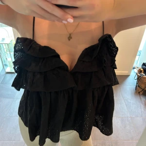 Svart volangig linnetopp med spets - Supersöt svart linnetopp med tunna axelband och flera lager volanger. Toppen har broderade hål och spetsdetaljer som ger en romantisk vibe. Perfekt för varma dagar när du vill ha något luftigt och snyggt.