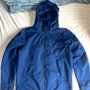 Blå vindjacka från Polo Ralph Lauren - Snygg marinblå vindjacka från Polo Ralph Lauren med huva och dragkedja framtill. Jackan har meshfoder, två fickor med dragkedja och klassisk röd logga broderad på bröstet. Justerbar snörning i huvan och sportig look, perfekt för blåsiga dagar.