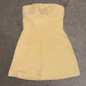Gul axelbandslös kortklänning - Snygg gul axelbandslös kortklänning med markerad byst och två fickor framtill med vita detaljer. Aldrig använd. Storlek 38, säljs för 200 kr