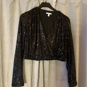 Svart paljettopp från H&M, croppad - Glittrig svart croppad topp från H&M med paljetter över hela ytan. Toppen har omlott-skärning framtill, v-ringning och långa ärmar. Perfekt för dig som vill sticka ut med en festlig look och gillar att glänsa.
