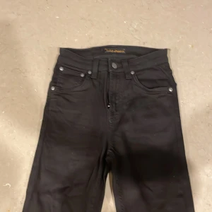 Svarta jeans från Nudie Jeans, W26 L30 - Svarta jeans från Nudie Jeans med klassisk femficksdesign och dragkedjegylf. Jeansen har raka ben och subtila detaljer på bakfickorna. Tillverkade i slitstarkt bomullstyg med en snygg, mörk finish. Perfekta för dig som gillar stilrena och tidlösa jeans.