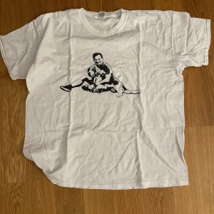 Vit Ulf Kristersson t-shirt - Vit t-shirt i bomull med ett stort svart tryck på bröstet som föreställer Ulf Kristersson som sitter och kramar en hund. Klassisk passform och rund halsringning. Perfekt för dig som gillar unika och personliga motiv.