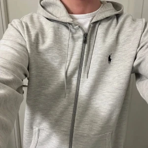Grå hoodie från Polo Ralph Lauren - Snygg grå hoodie från Polo Ralph Lauren med dragkedja framtill och klassisk huva. Tröjan har två fickor, snörning i huvan och den ikoniska svarta loggan broderad på bröstet. Perfekt för en chill och stilren look. Passar L lika bra som den passar M!