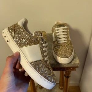Valentino glittriga - Säljer ett par Valentino Rockstud sneakers i guldglittrigt material med vita skinnpaneler och vita snören.storlek 36 men passar absolut 37 Skorna har ikoniska nitar på hälen och detaljer i guld. Snygg och lyxig look som verkligen sticker ut. Perfekta för dig som vill ha något extra.